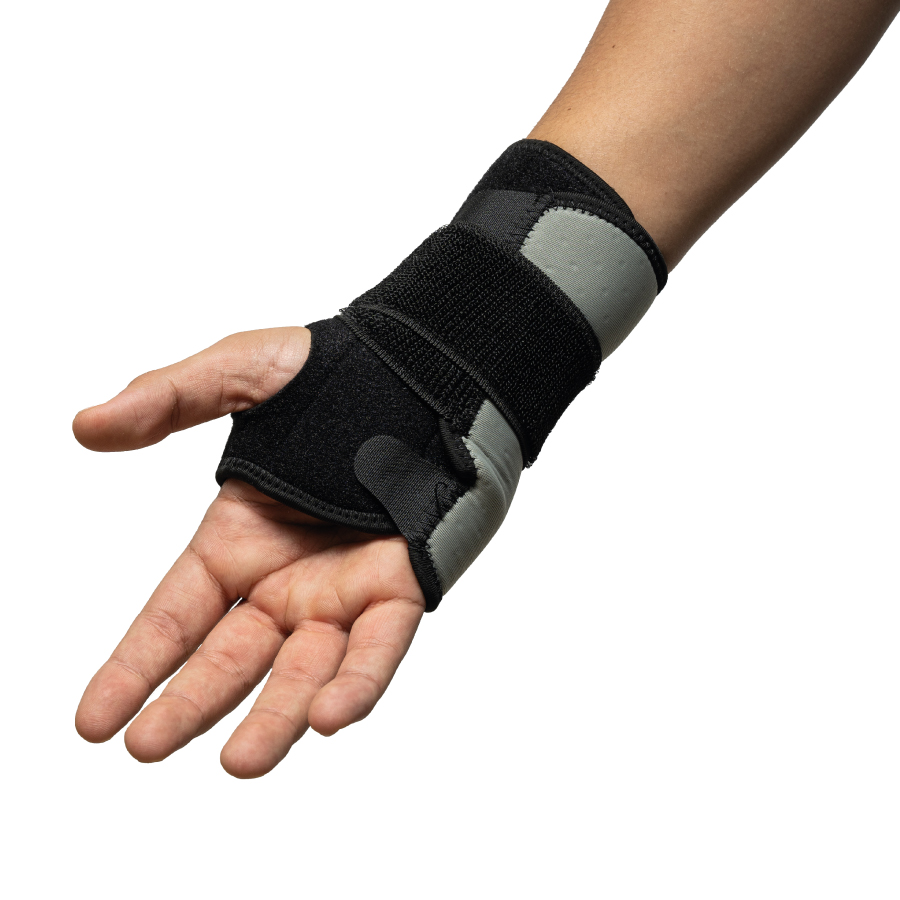 Brace de muñeca ambidiestro GMD, puesto en la mano derecha de una persona. De color negro y gris, con correa de velcro negra que rodea la muñeca, cubre desde la muñeca hasta la palma de la mano, dejando los dedos libres y con abertura para el pulgar.. Haga clic para ampliar la imagen