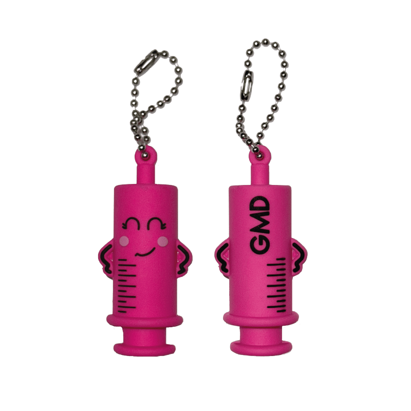 Rompe ampollas color rosado, tipo jeringa, sonriente y manos a los lados, detalles en color negro y, en la parte posterior, líneas de medición marcadas en el cuerpo con el texto “GMD”; con cadena tipo llevero de bolas plateada.. Haga clic para ampliar la imagen