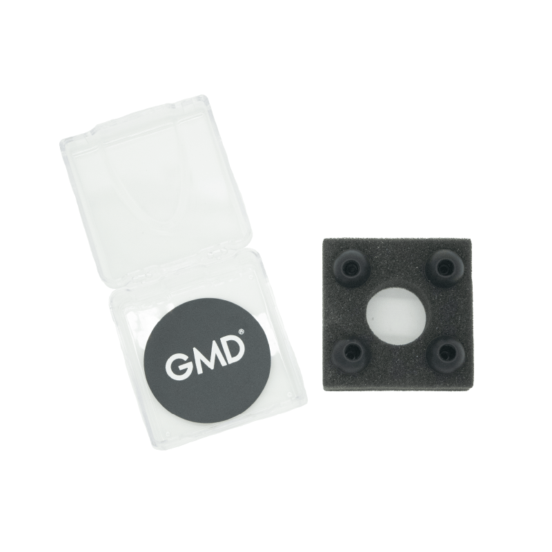 Set de accesorios para OIS 500, caja transparente abierta con membrana circular negra y logo GMD impreso en blanco. A su lado, espuma negra con cuatro olivas de goma negras organizadas alrededor de una abertura circular central.. Haga clic para ampliar la imagen